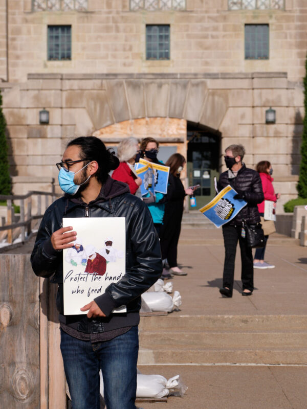 Community members stand at the east entrance of the Nebraska State Capitol, holding signs encouraging protections for essential workers.