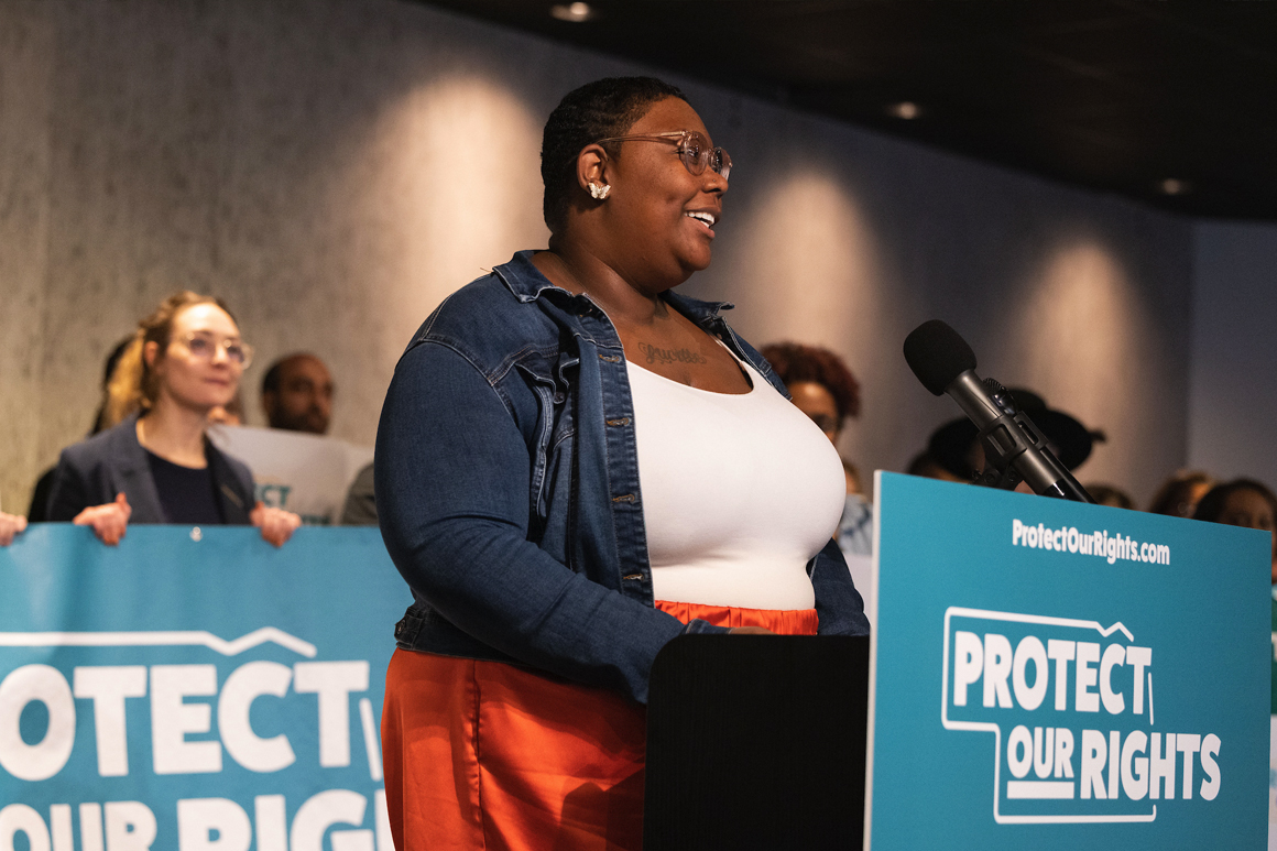 Jasmine Smith, a sworn sponsor of the Protect Our Rights initiative, speaks at the campaign launch. Jasmine is a black woman with short-cropped hair, wearing a blue jacket and white blouse.