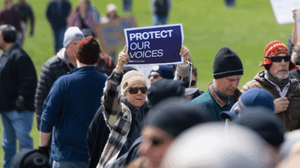A rally attendee holds up a sign reading "protect our voices"