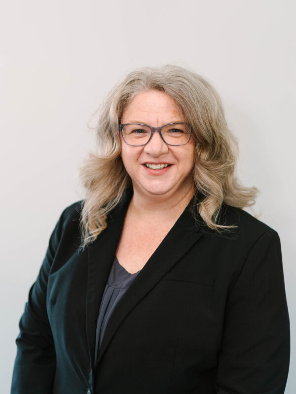 A white woman with glasses and a black blazer smiles, posing for a photograph.