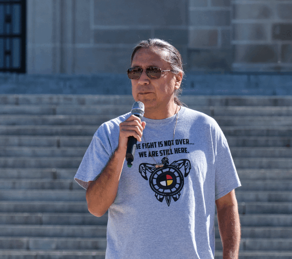 Kevin Abourezk speaks in front of the Nebraska State Capitol wearing a shirt that reads "The fight is not over. We are still here."