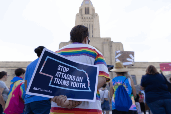 A rally attendee holds a sign showing the outline of Nebraska in light blue, white and pink. It reads "Stop attacks on trans youth."