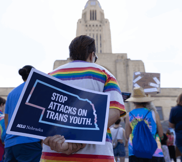 A rally attendee holds a sign showing the outline of Nebraska in light blue, white and pink. It reads "Stop attacks on trans youth."