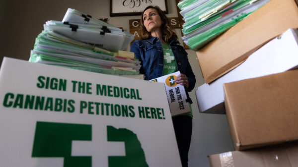 A woman in a denim jacket and green shirt looks into the distance while holding a petition. In the foreground, there is a sign that reads "Sign the Medical Cannabis Petitions Here!"