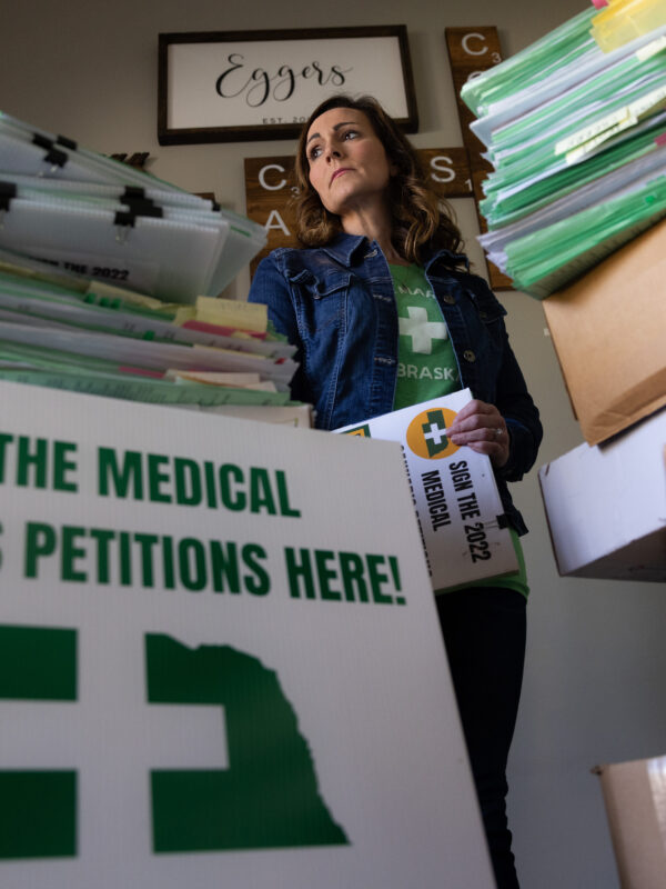 A woman in a denim jacket and green shirt looks into the distance while holding a petition. In the foreground, there is a sign that reads "Sign the Medical Cannabis Petitions Here!"