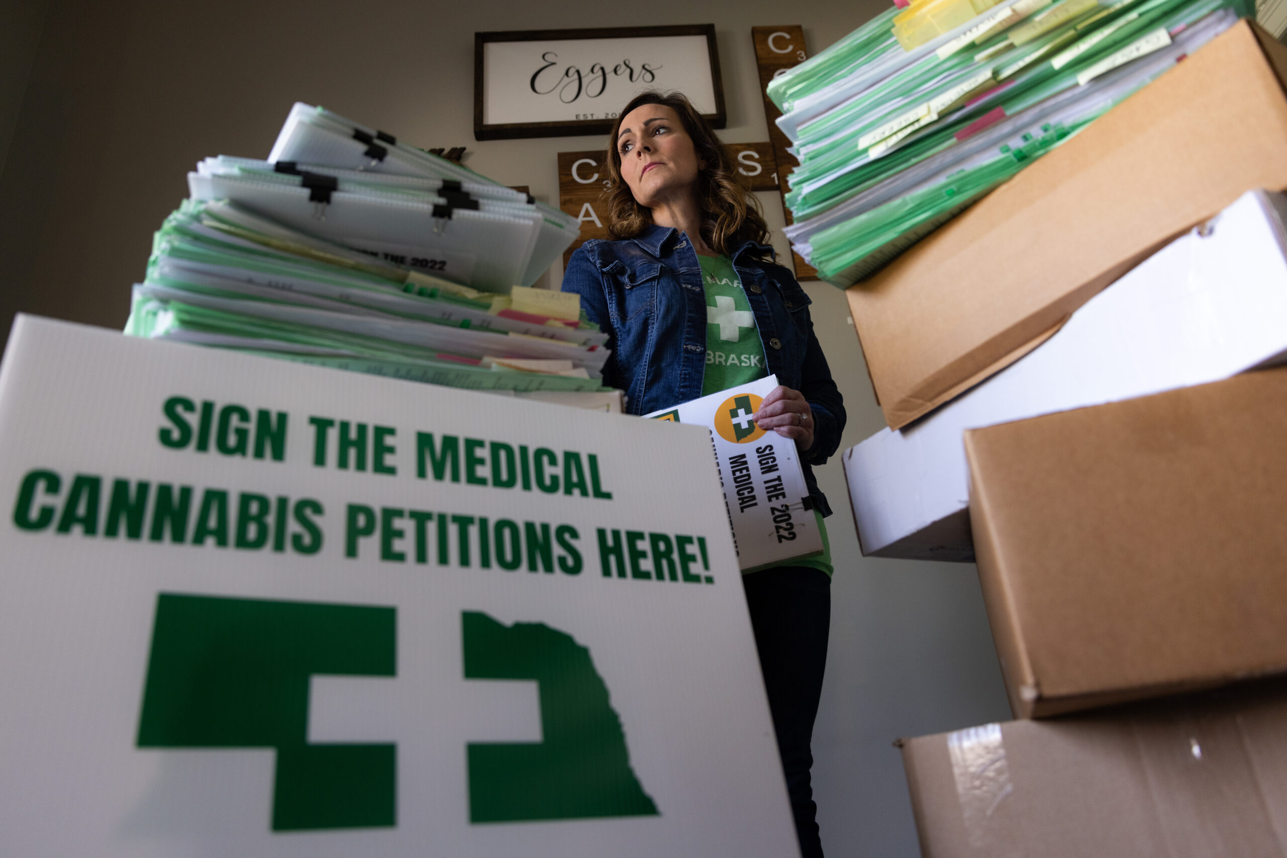 A woman in a denim jacket and green shirt looks into the distance while holding a petition. In the foreground, there is a sign that reads "Sign the Medical Cannabis Petitions Here!"