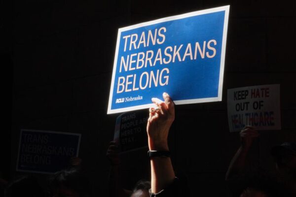 A blue sign with pink words reading "Trans Nebraskans Belong" being held by one hand against a dark background.