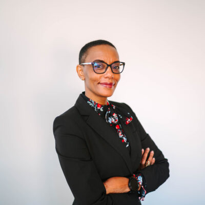 Jamel Connor standing in front of a white background. She has short hair, black rimmed glasses, a black coat and her arms crossed.