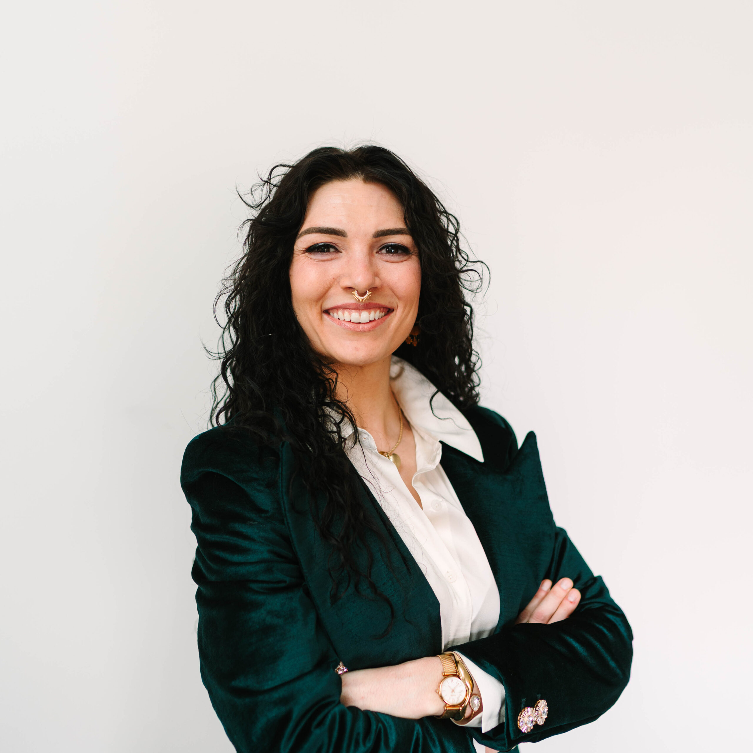 Cassy Ross standing in front of a white wall with a emeral green blazer and a white button up. She has long, curly black hair.