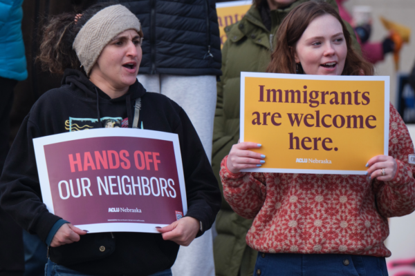 Two protest attendees hold ACLU signs related to immigrants' rights. One reads "Hands off our neighbors." The other reads "Immigrants are welcome here."