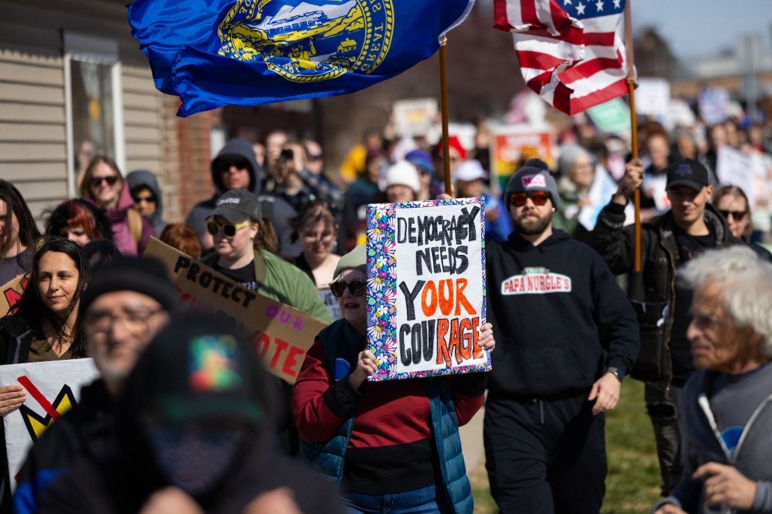Protesters in Omaha walk along a sidewalk. One holds a sign reading "Democracy needs our courage." Different colors call out the letters writing "our rage."