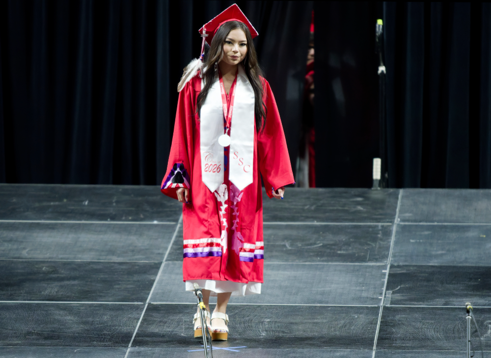 Tiala Luong walks to receive her diploma in a ribboned gown and wearing an eagle feather.