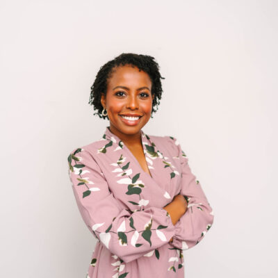 Yvonne Kaburu, standing in front of a white while with her arms crossed. She is wearing a pink dress with white flowers and has short curly hair.