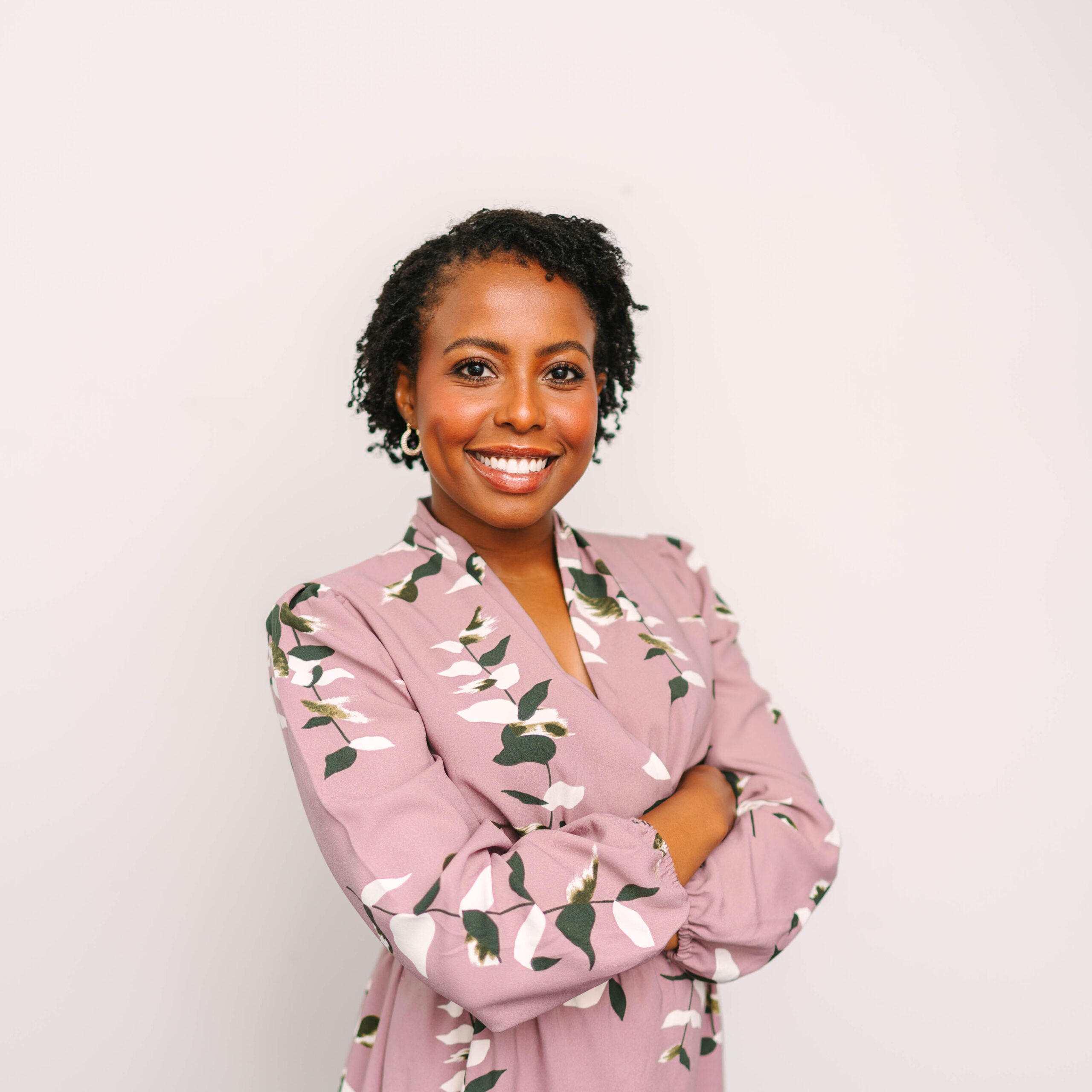 Yvonne Kaburu, standing in front of a white while with her arms crossed. She is wearing a pink dress with white flowers and has short curly hair.