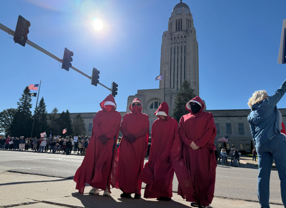 A group of women in red robes and white bonnets, inspired by Margaret Atwood's Handmaid's Tale, stand outside the Nebraska State Capitol.