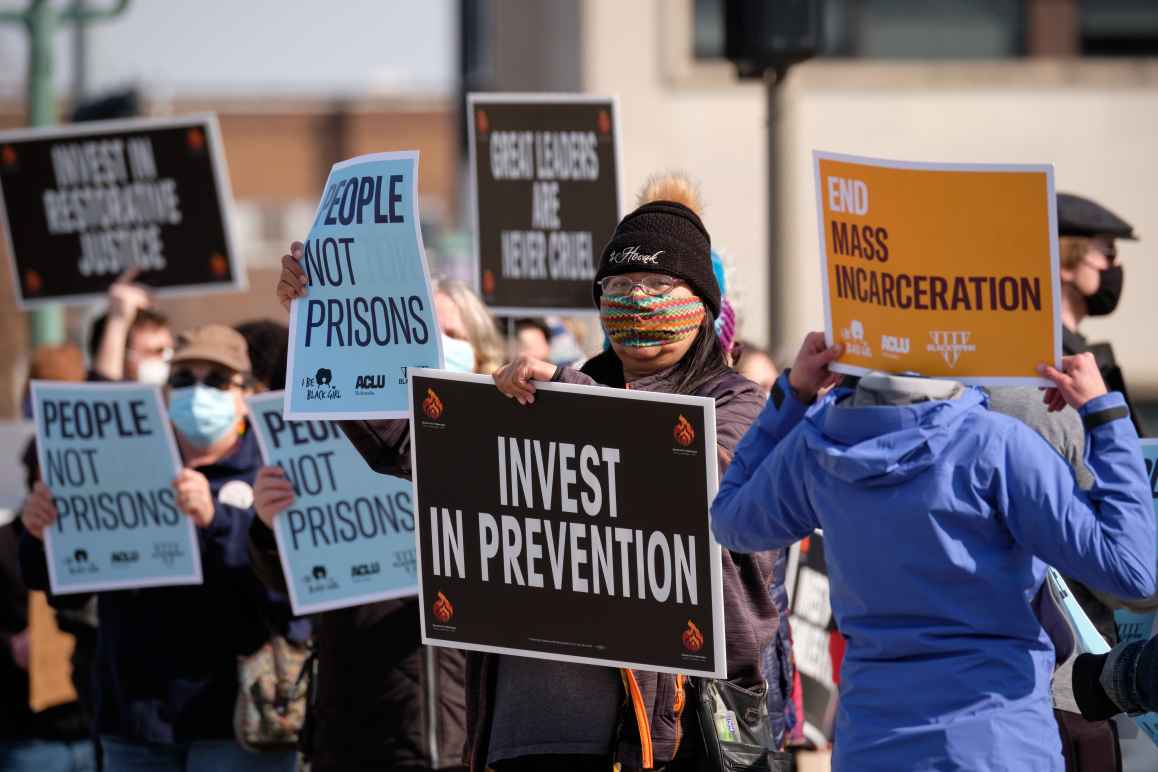 Protesters stand together, holding signs protesting the new prison. One poster reads "Invest in prevention."