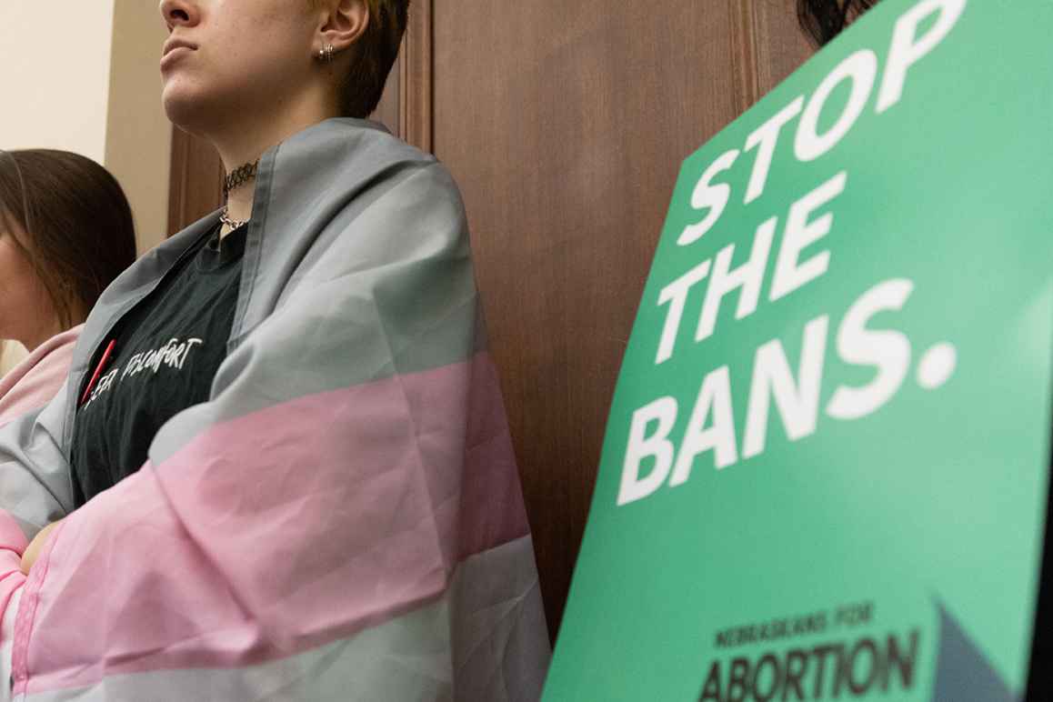 A Nebraskan wrapped in the trans flag listens to testimony during a committee hearing for the near-total abortion ban.