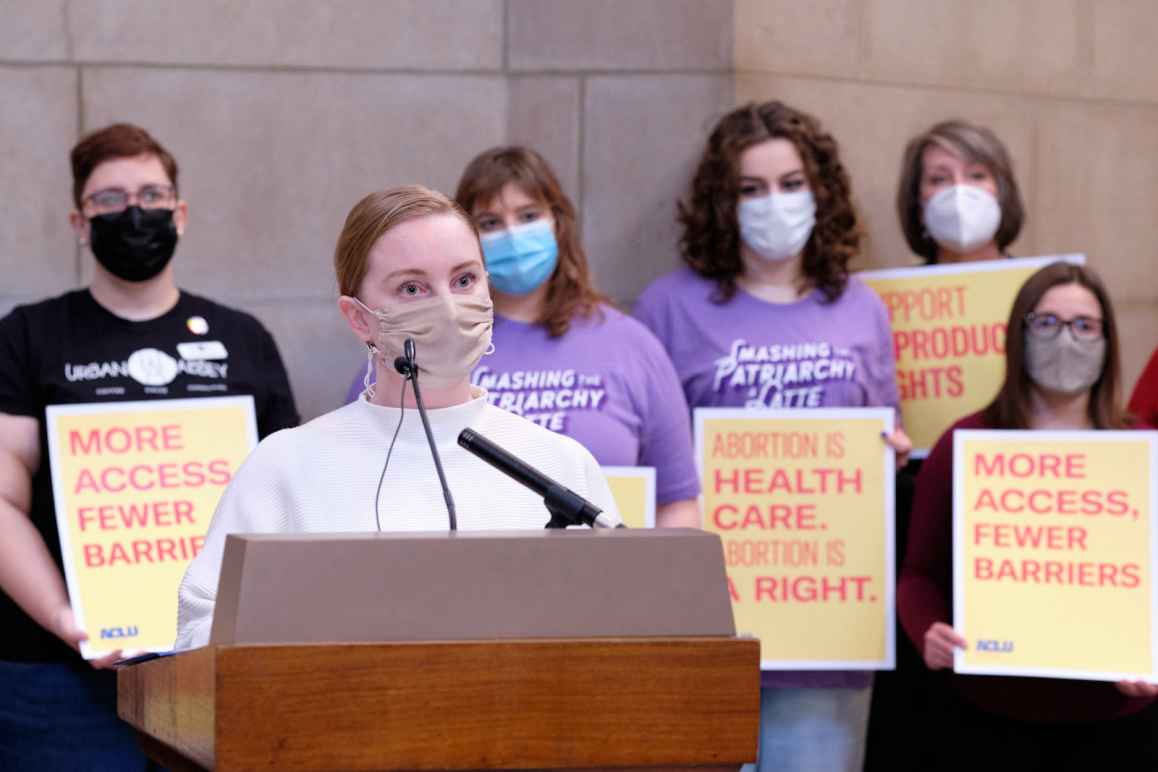 Senator Megan Hunt speaks in front of a group holding signs reading "Abortion is Health Care Abortion is a Right" and "More access, fewer barriers"