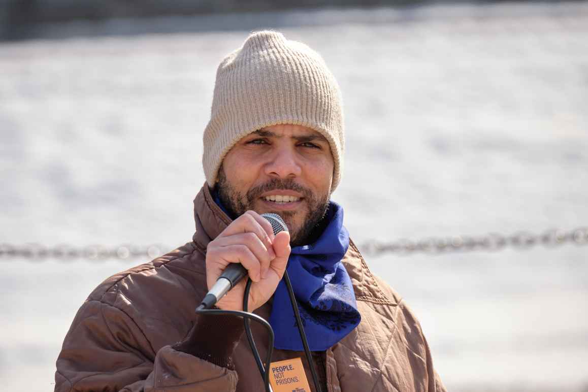 Jason Witmer, a brown-skinned African American man, wears a tan stocking cap and a state-issued brown jacket adorned with a button that boldly declares "People Not Prison." Jason stands confidently poised, holding a microphone to his lips.