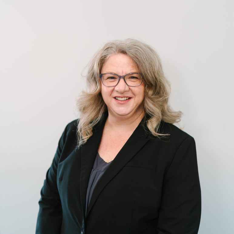 A photo of a smiling white woman in a black blazer with glasses and long hair.