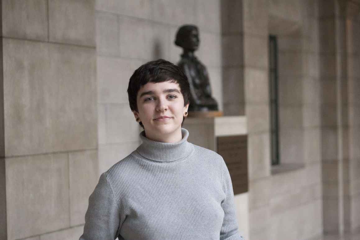 Mar Lee wears a light grey turtleneck, standing in front of a bust