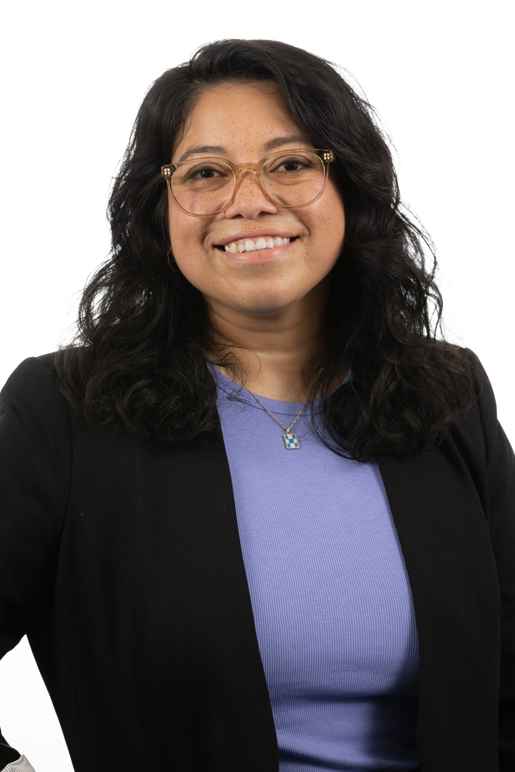 This image of Rose Godinez shows a smiling woman with tan skin tone and slightly longer than shoulder-length black wavy hair. Rose is wearing a light blue top with a black suit jacket, round light brown glasses, and a necklace.