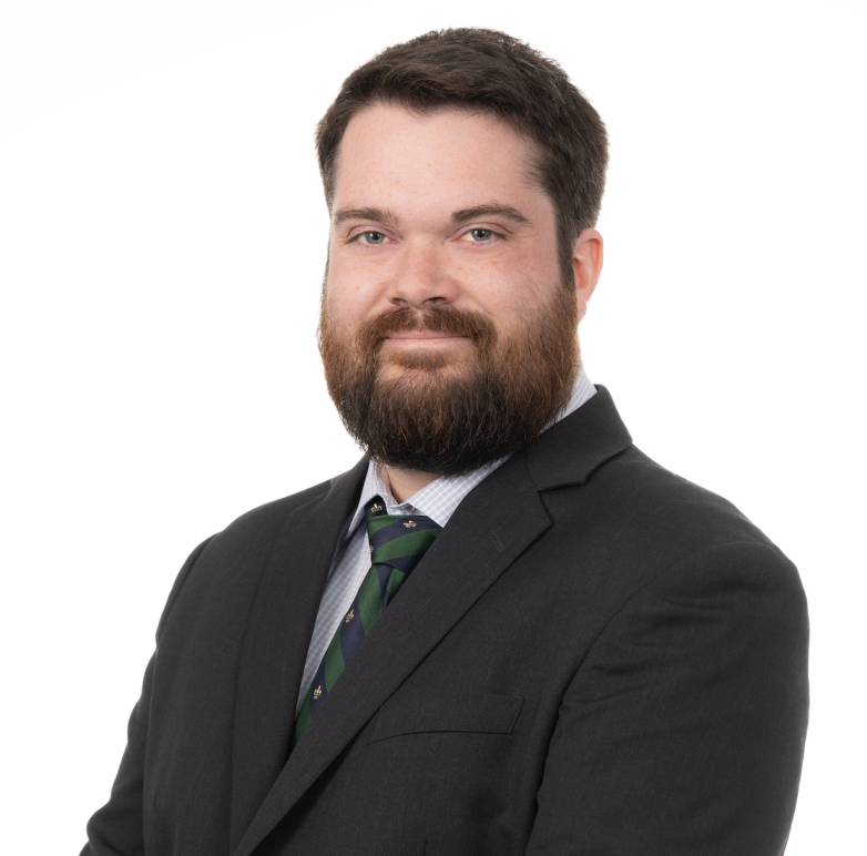This image of Dylan Severino shows a smiling white man with short, dark hair and a trimmed full beard. He is wearing a black suit and a striped blue-green tie.