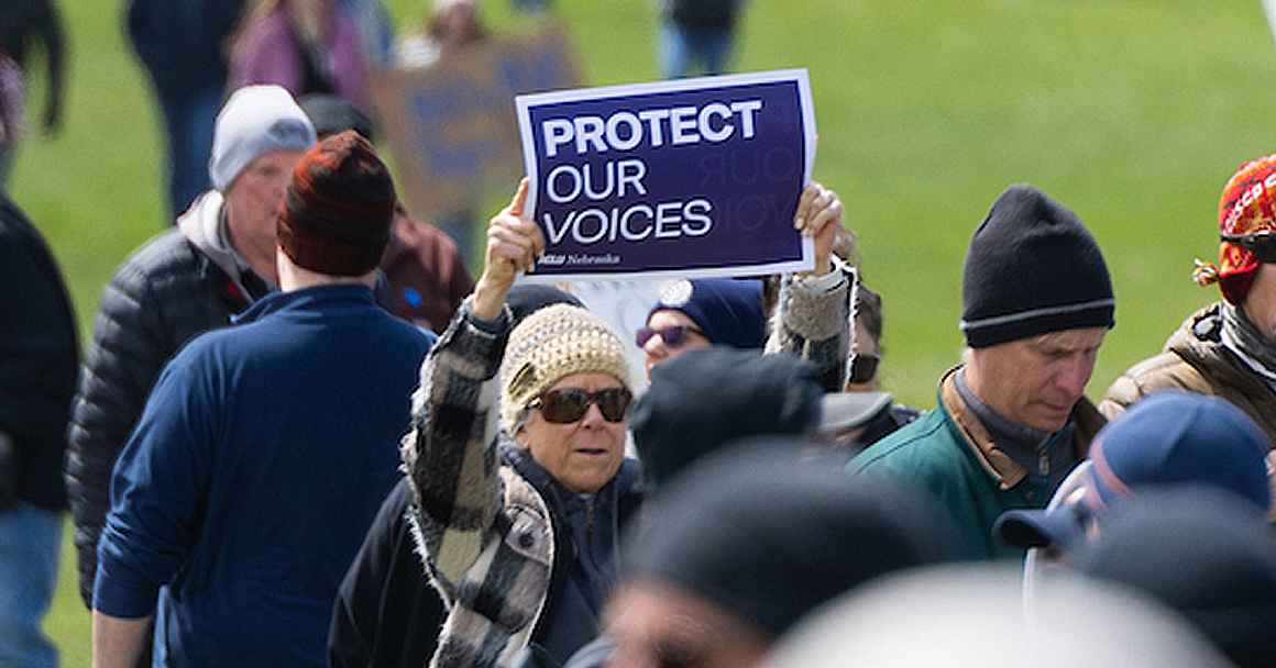 A rally attendee holds a purple and white ACLU of Nebraska sign reading "Protect our voices"
